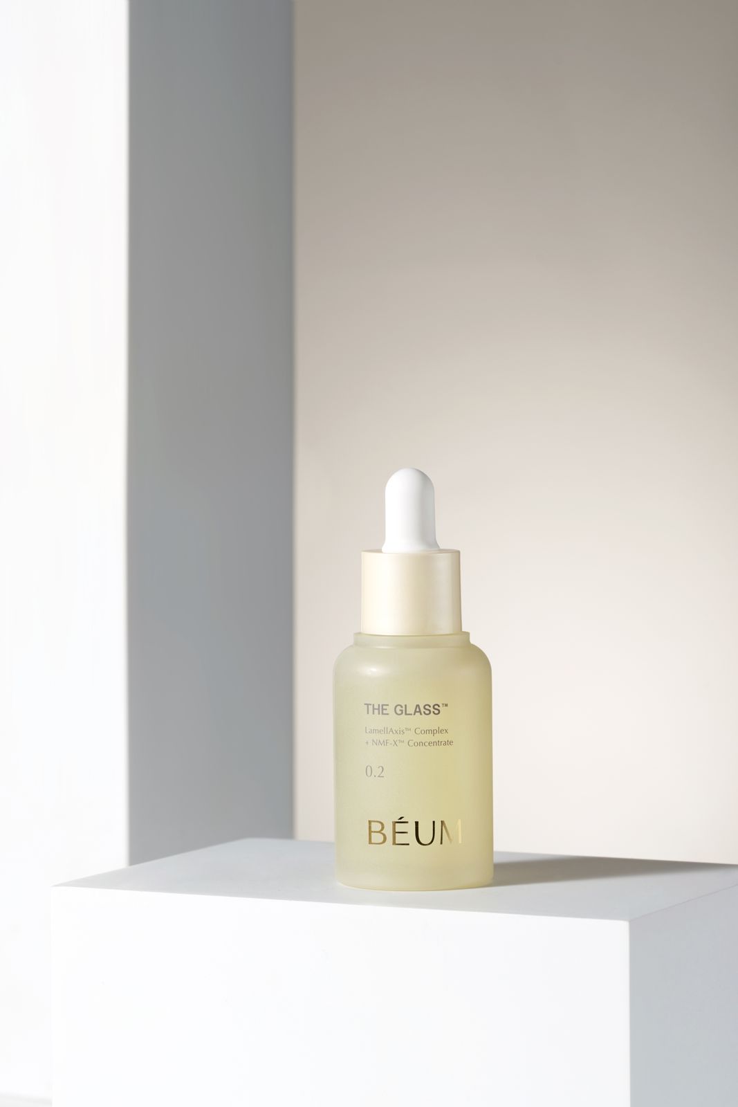 BEUM · 더글래스 스킨케어 4종 — 0.2 세럼 병을 화이트 블록 모서리 위에 세운 단독컷, 웜톤 그라데이션 배경