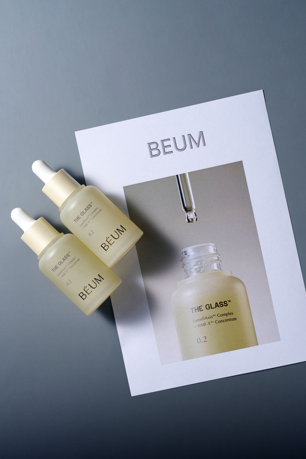 BEUM · 더글래스 스킨케어 4종 — 다크 블루 배경 위에 브랜드 포스터를 깔고 0.2 세럼 병 2개를 올린 연출컷