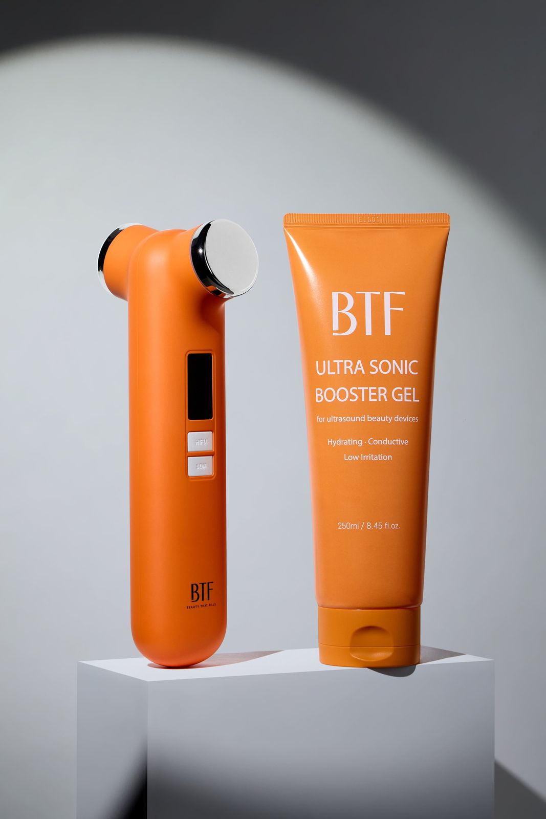 BTF · 부스터젤 — 디바이스와 부스터젤을 큐브 좌대 위에 나란히 놓은 컷, 원형 스포트라이트 배경