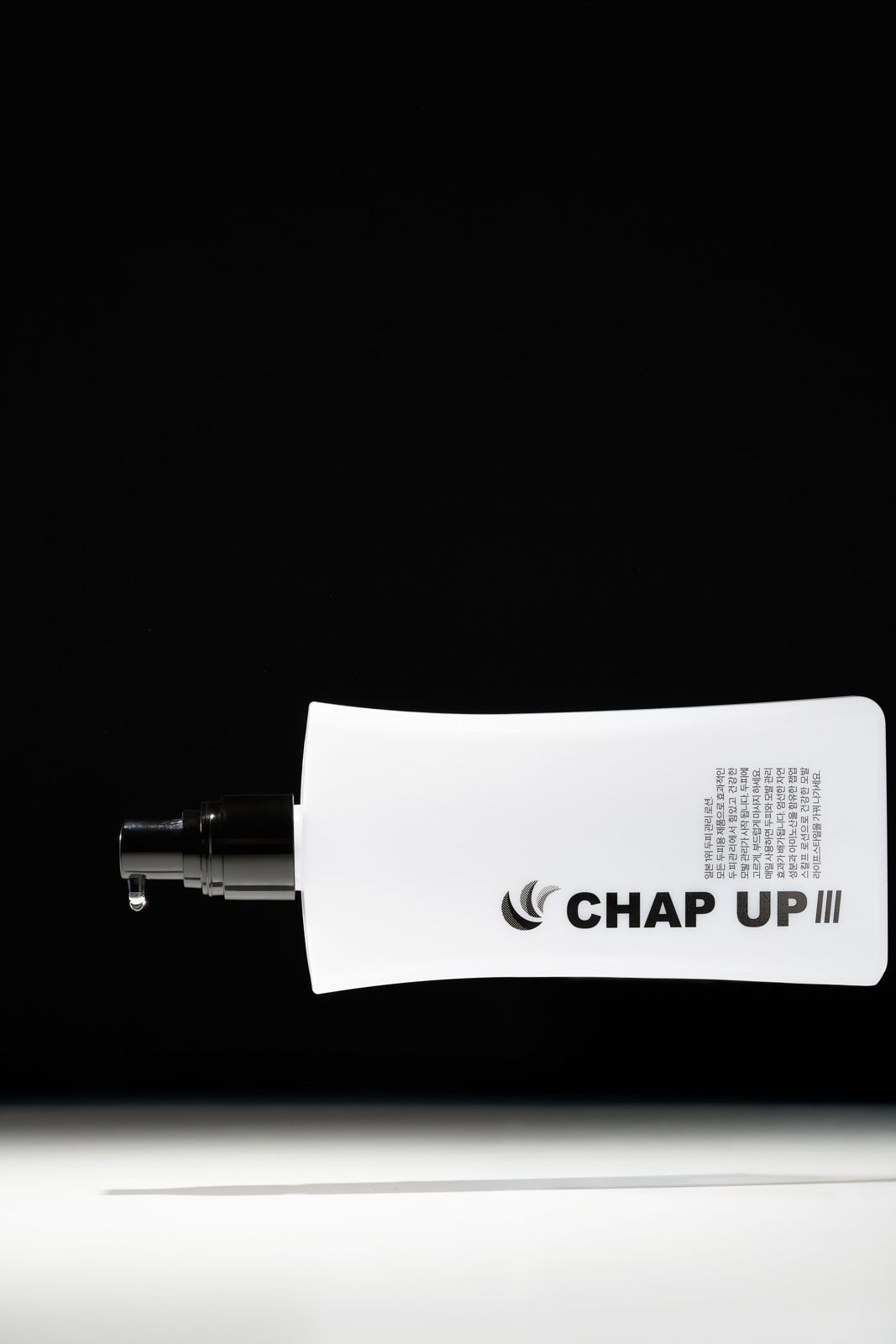 CHAPUP · 두피로션 — 노즐 끝에 맺힌 제형 방울을 보여주는 옆면 클로즈업, 화이트 스포트라이트 바닥, 블랙 배경