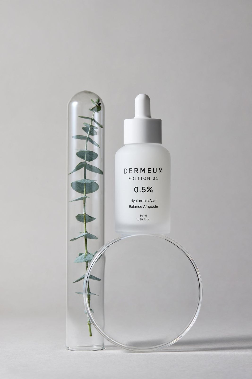 DERMEUM · 앰플 — 유칼립투스 잎이 든 유리관과 제품을 나란히 배치한 컷, 그레이 배경