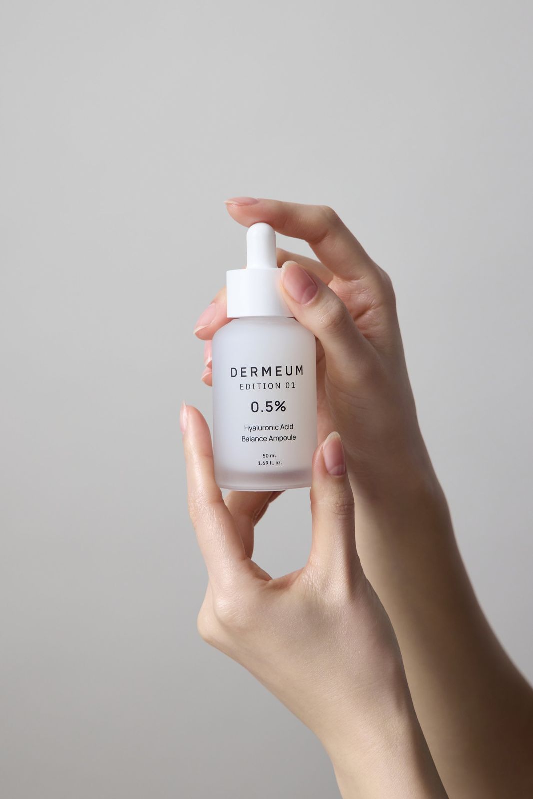 DERMEUM · 앰플 — 손끝으로 드로퍼 뚜껑을 잡고 제품을 들어 올린 핸드모델 컷, 그레이 배경