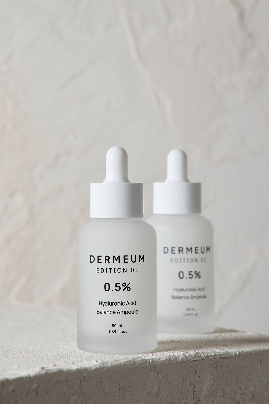 DERMEUM · 앰플 — 제품 2개를 텍스처 있는 배경 위에 나란히 세운 컷, 베이지 배경
