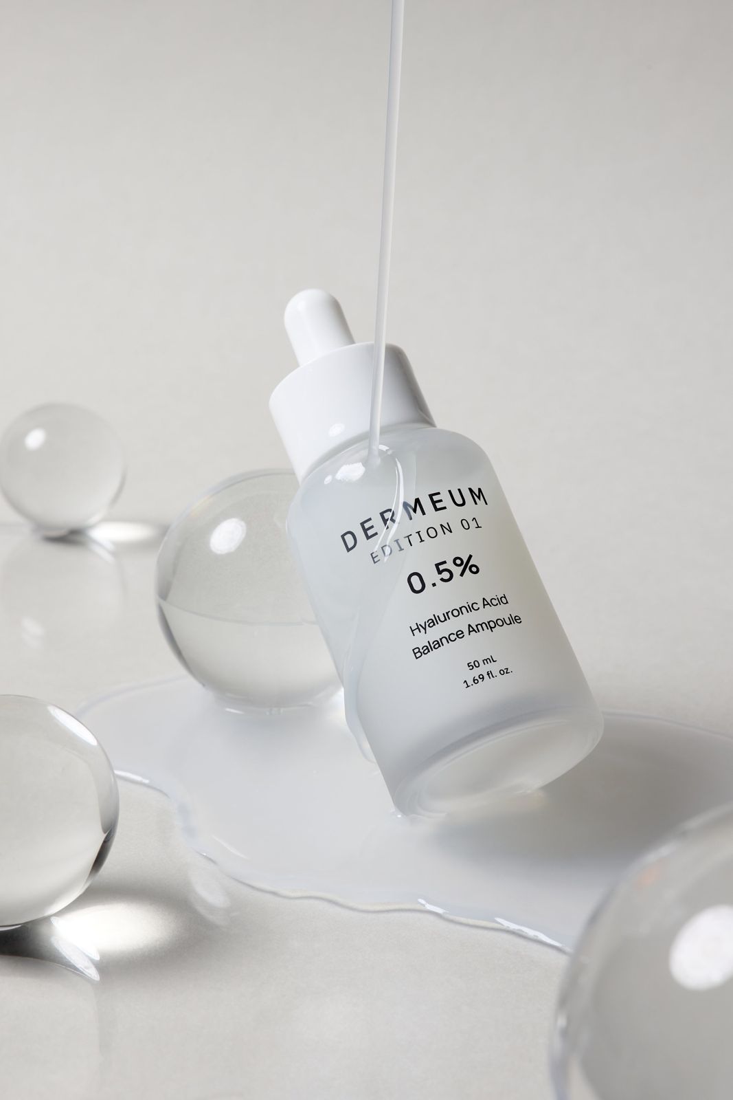DERMEUM · 앰플 — 유리구슬 소품 사이로 제품에서 제형이 흘러내리는 텍스처컷, 화이트 배경