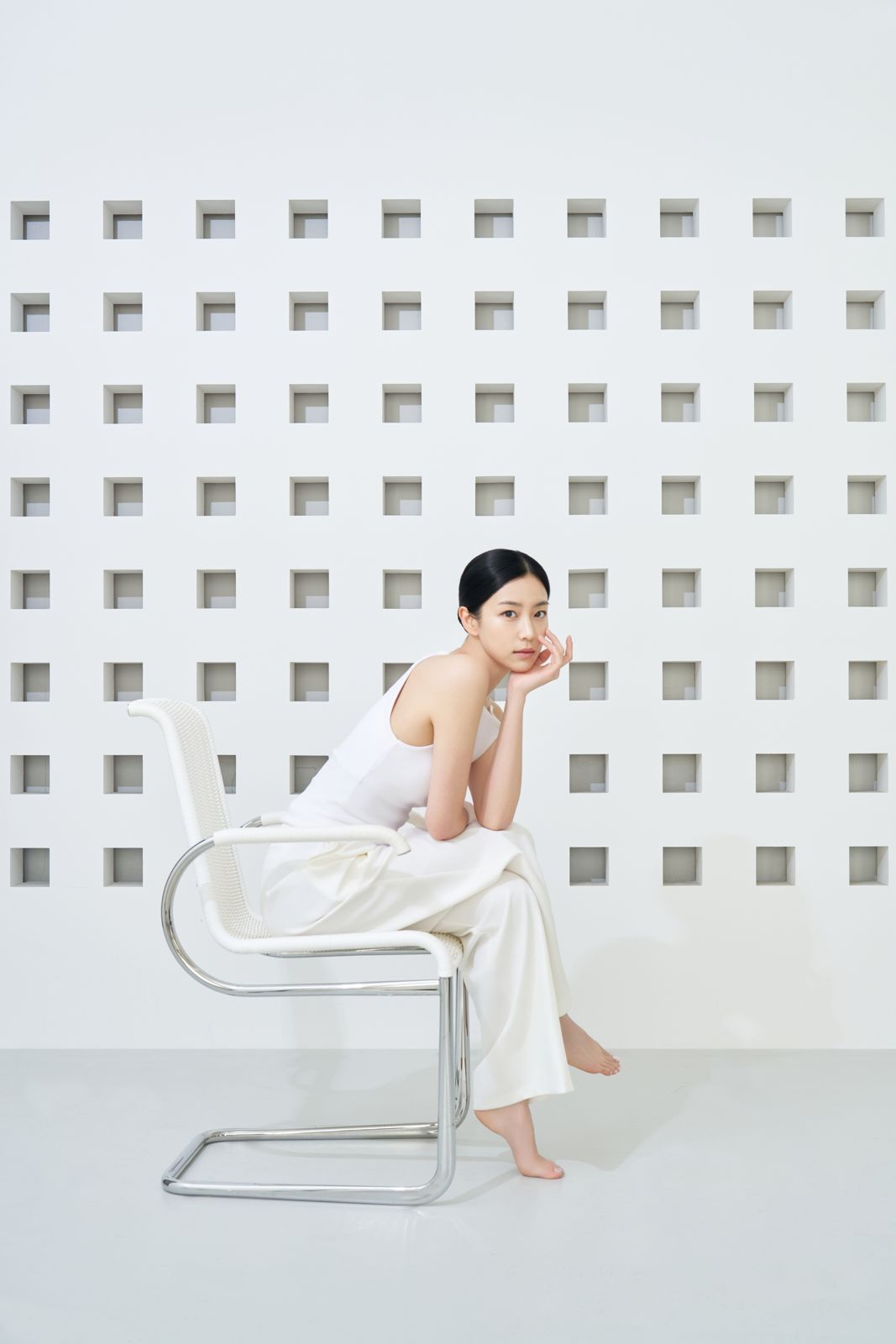DEWISL · 모델촬영 — 화이트 디자인 체어에 앉아 턱을 괸 모델컷, 격자무늬 벽면 배경