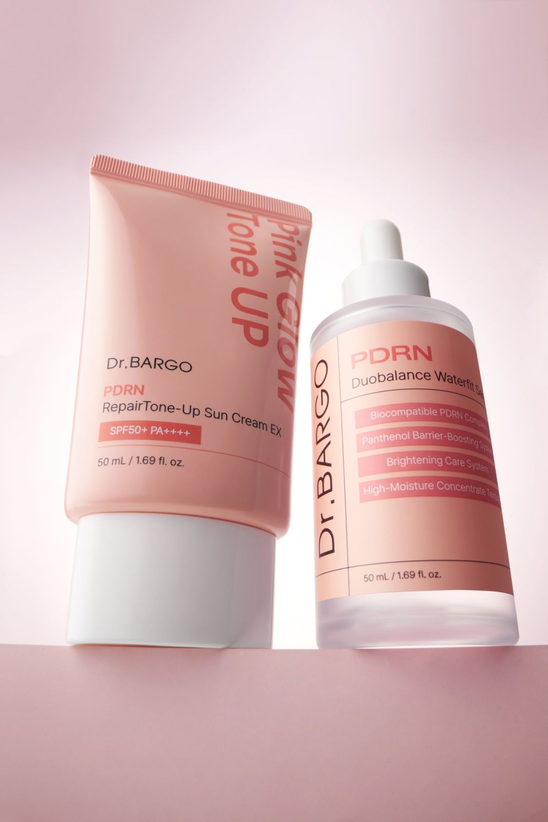 Dr.BARGO · 선크림 — 선크림 튜브를 기울여 세럼 드로퍼 병과 나란히 배치한 크로스 프로덕트 연출컷, 핑크 배경