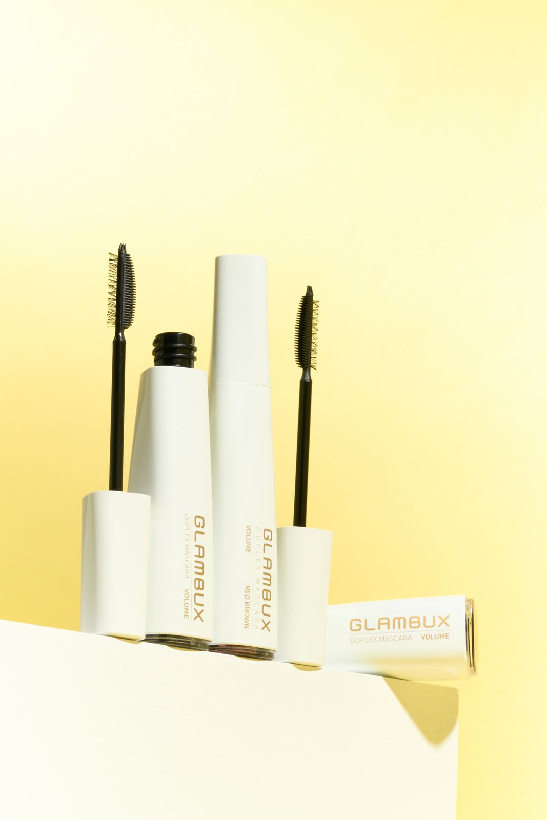 GLAMBUX · 마스카라 — 화이트 좌대 모서리에 튜브 여러 개를 세우거나 걸쳐 놓은 컷, 옐로우 배경