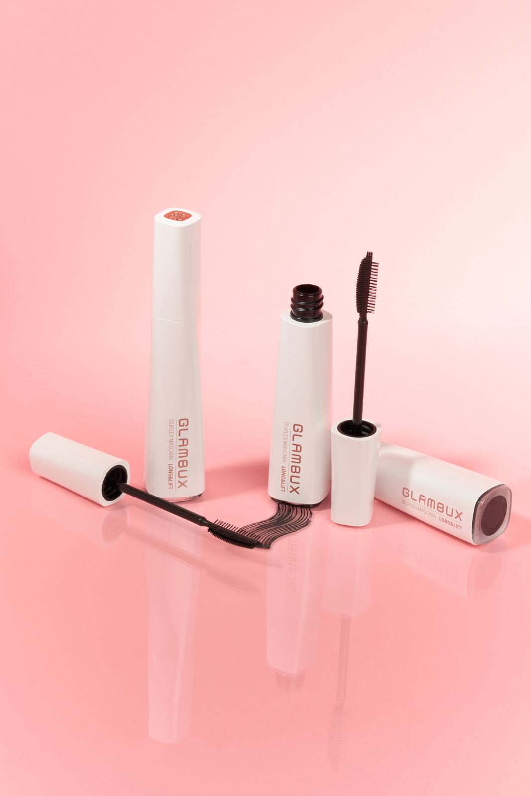 GLAMBUX · 마스카라 — 반사 바닥 위 핑크 배경, 튜브 3개와 브러시, 제형 스와치를 함께 배치한 그룹컷