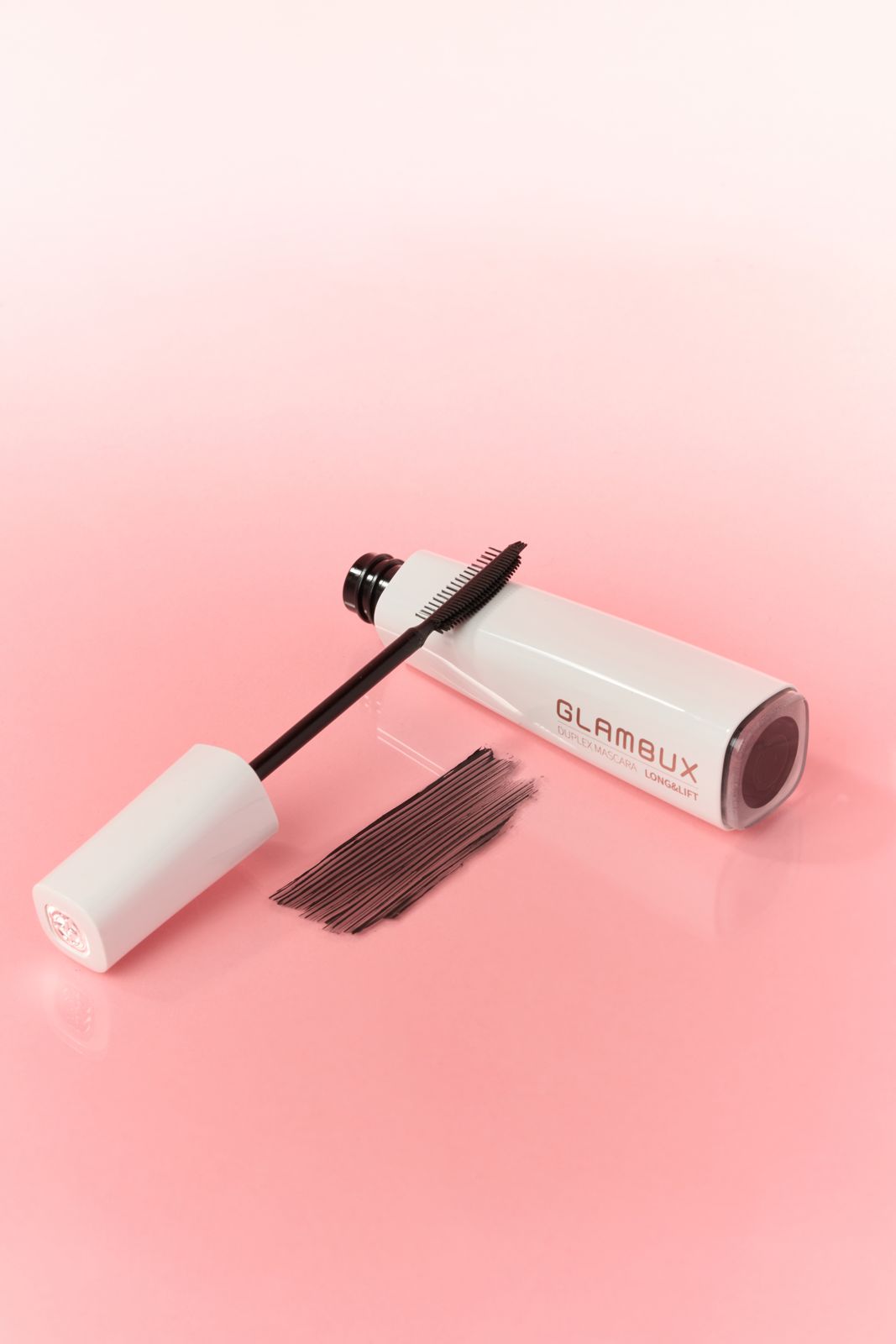 GLAMBUX · 마스카라 — 핑크 배경, 튜브를 투명 받침에 살짝 기울여 세우고 브러시와 제형 스와치를 함께 놓은 컷