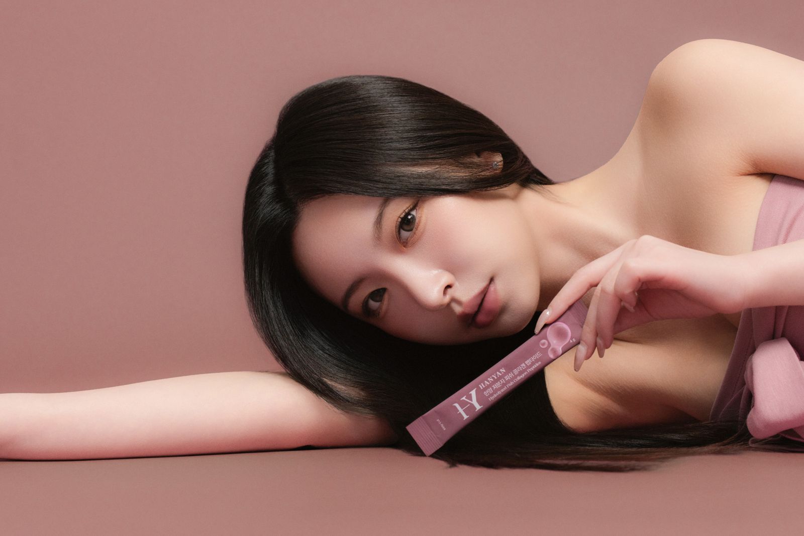 HANYAN · 건강기능식품 (모델컷) — 누워서 사쉐 스틱을 손에 든 모델컷, 로즈핑크 배경
