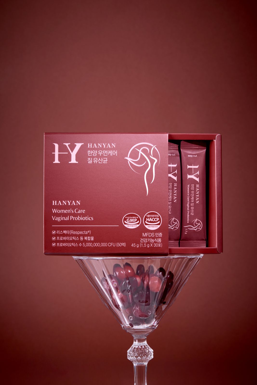HANYAN · 건강기능식품 — 버건디 배경, 붉은 알갱이가 담긴 칵테일 잔 위에 열린 박스를 균형 있게 얹은 연출컷