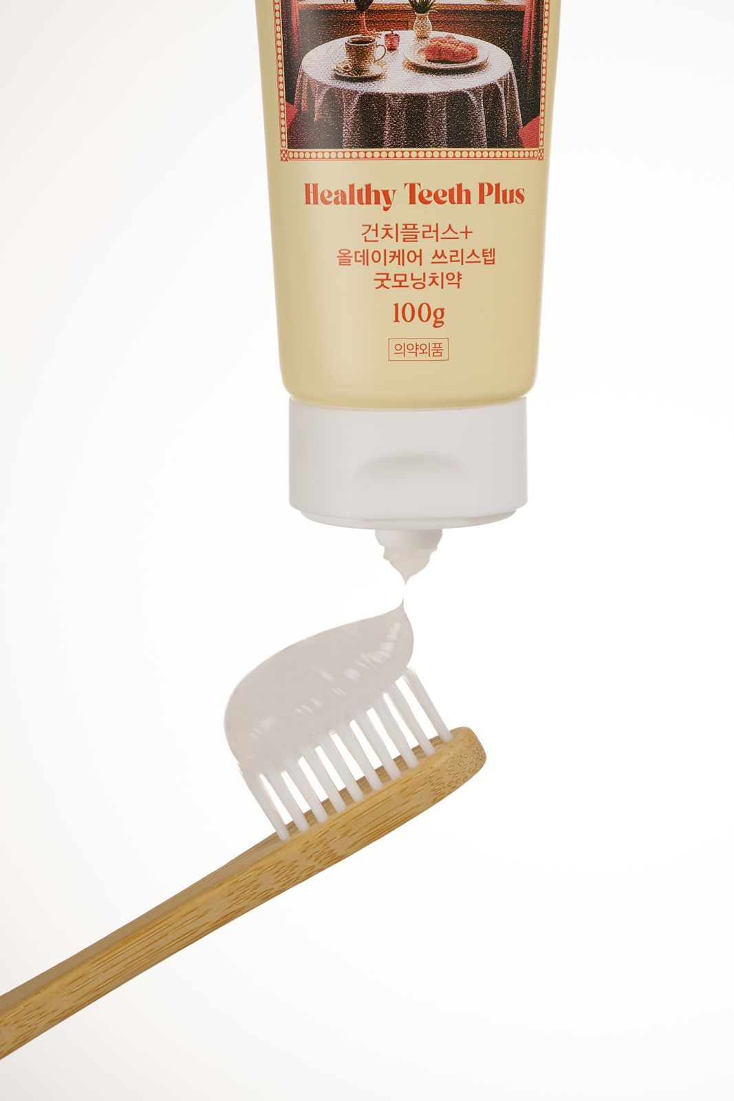 HEALTHY TEETH PLUS · 올데이케어 치약 — 굿모닝 치약을 나무 칫솔 위에 짜 올리는 클로즈업 연출컷, 베이지 배경