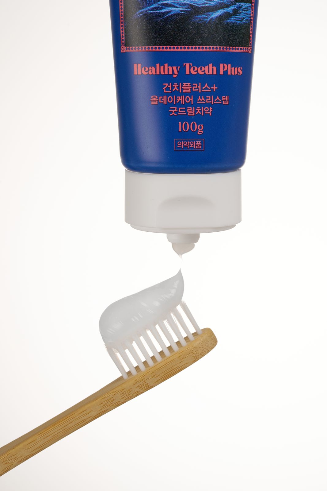 HEALTHY TEETH PLUS · 올데이케어 치약 — 굿드림 치약을 나무 칫솔 위에 짜 올리는 클로즈업 연출컷, 베이지 배경