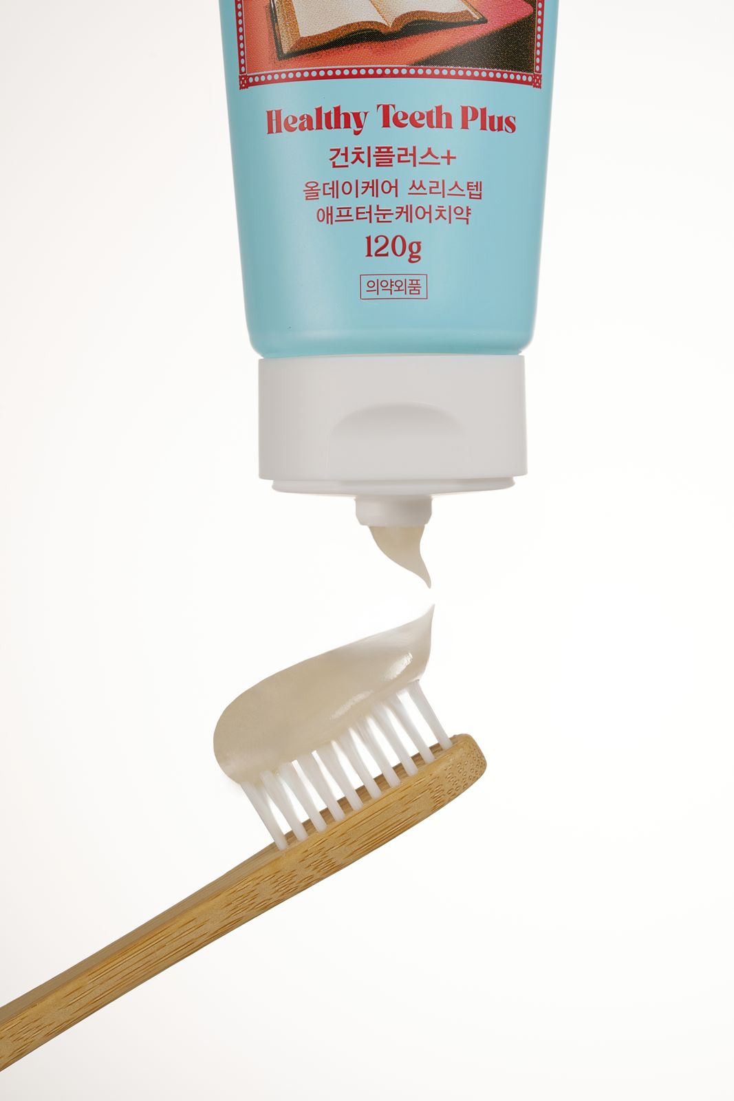 HEALTHY TEETH PLUS · 올데이케어 치약 — 애프터눈케어 치약을 나무 칫솔 위에 짜 올리는 클로즈업 연출컷, 베이지 배경
