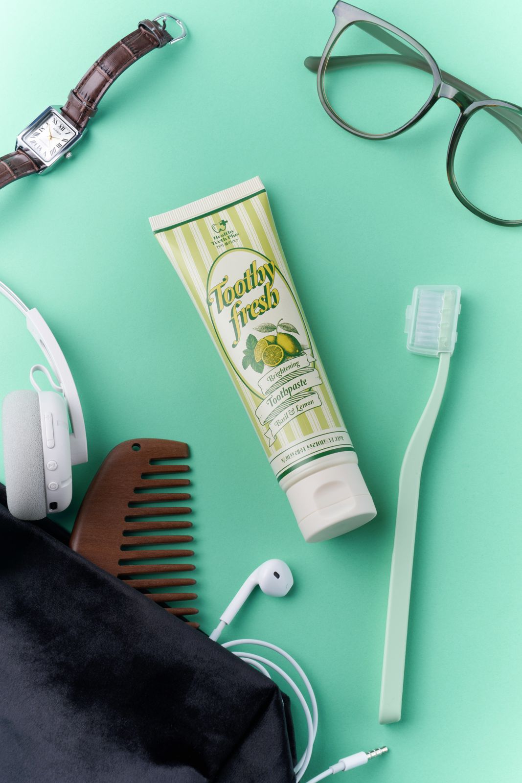 HEALTHY TEETH PLUS · 투씨프레시 브라이트닝 치약 — 민트 배경 위에 손목시계·안경·헤드폰·나무 빗·이어폰·파우치·칫솔 등 일상 소지품과 함께 놓은 플랫레이