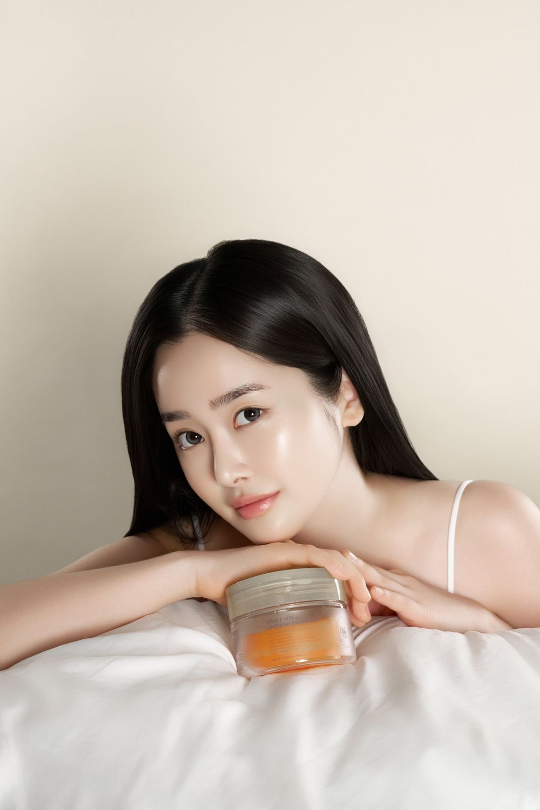 HEVEBLUE · 토너패드 — 하얀 침구에 기대어 제품 통을 턱 아래에 받쳐 든 모델 컷, 베이지 배경