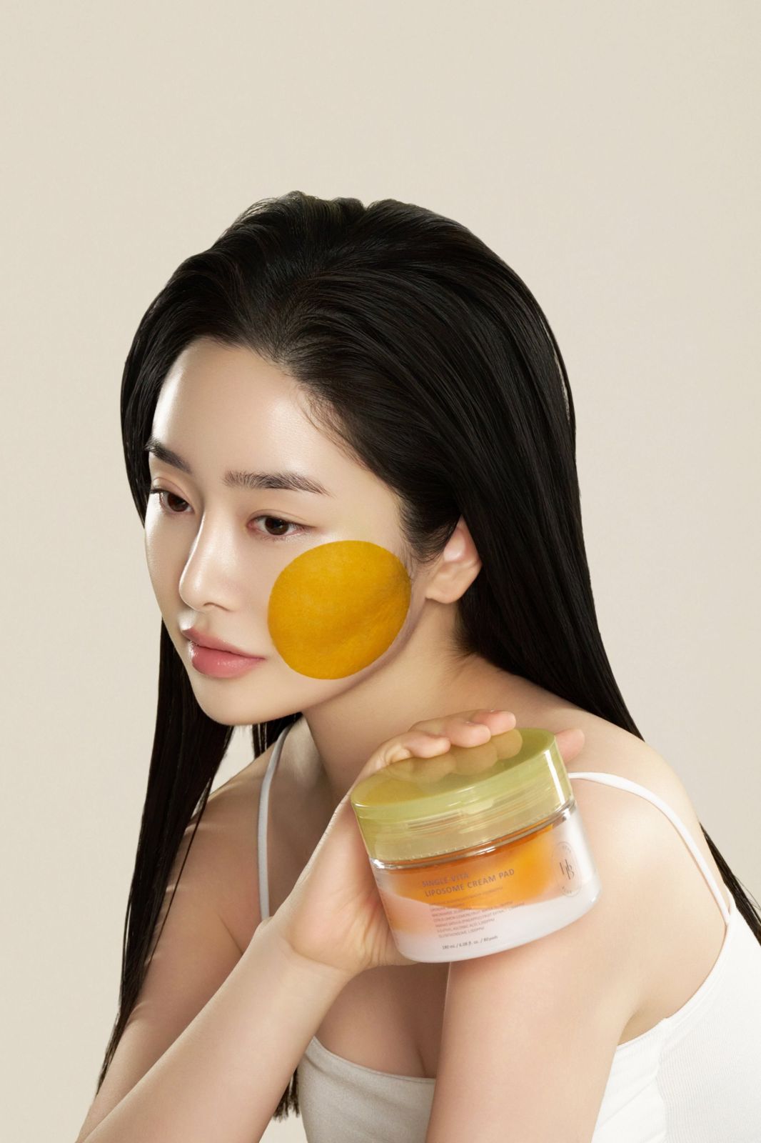 HEVEBLUE · 토너패드 — 패드를 뺨에 대고 제품 통을 함께 든 모델 뷰티컷, 베이지 배경