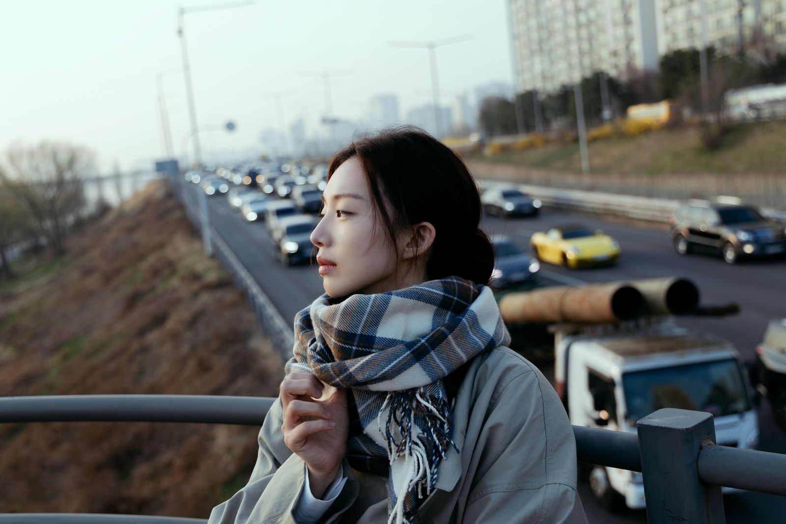 KINED · 모델촬영 — 고가도로 위에서 체크 목도리를 여미며 도로를 바라보는 컷(제품 미노출)