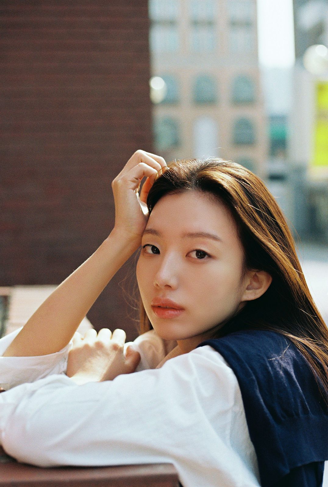 KINED · 모델촬영 — 도심 건물 배경, 네이비 베스트 착장의 얼굴 클로즈업 필름톤 컷(제품 미노출)