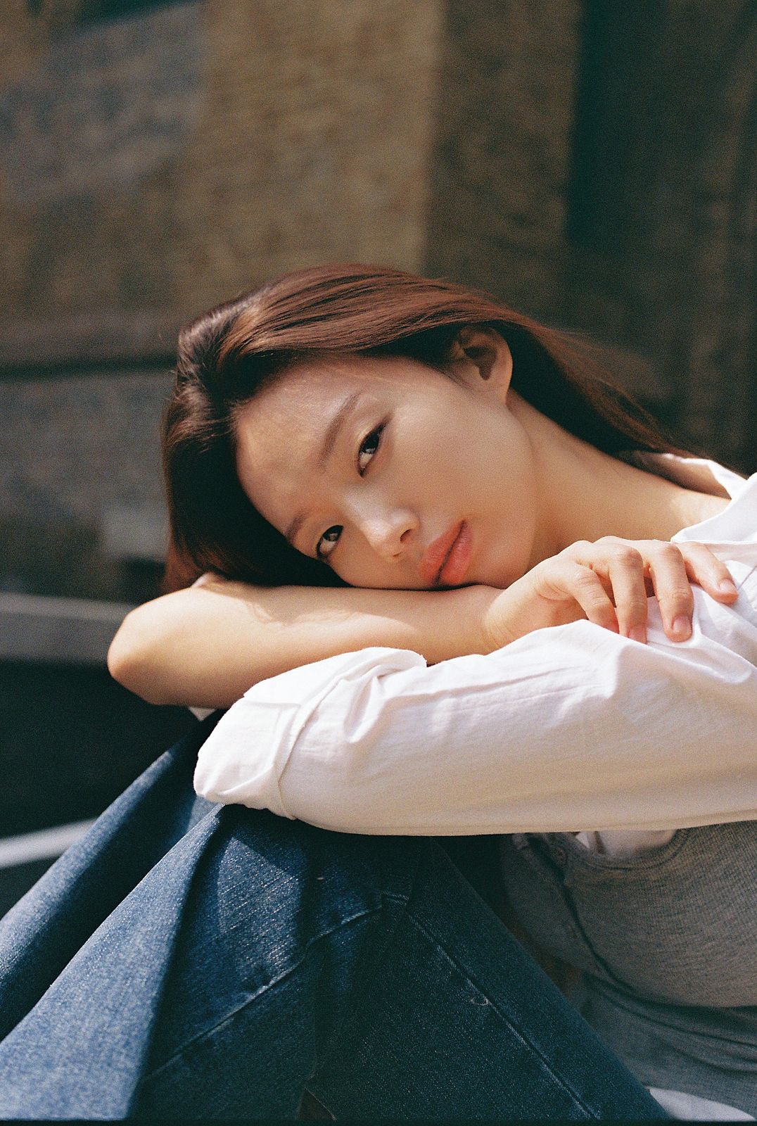 KINED · 모델촬영 — 무릎에 팔을 얹고 기댄 채 정면을 응시하는 필름톤 얼굴 클로즈업 컷(제품 미노출)