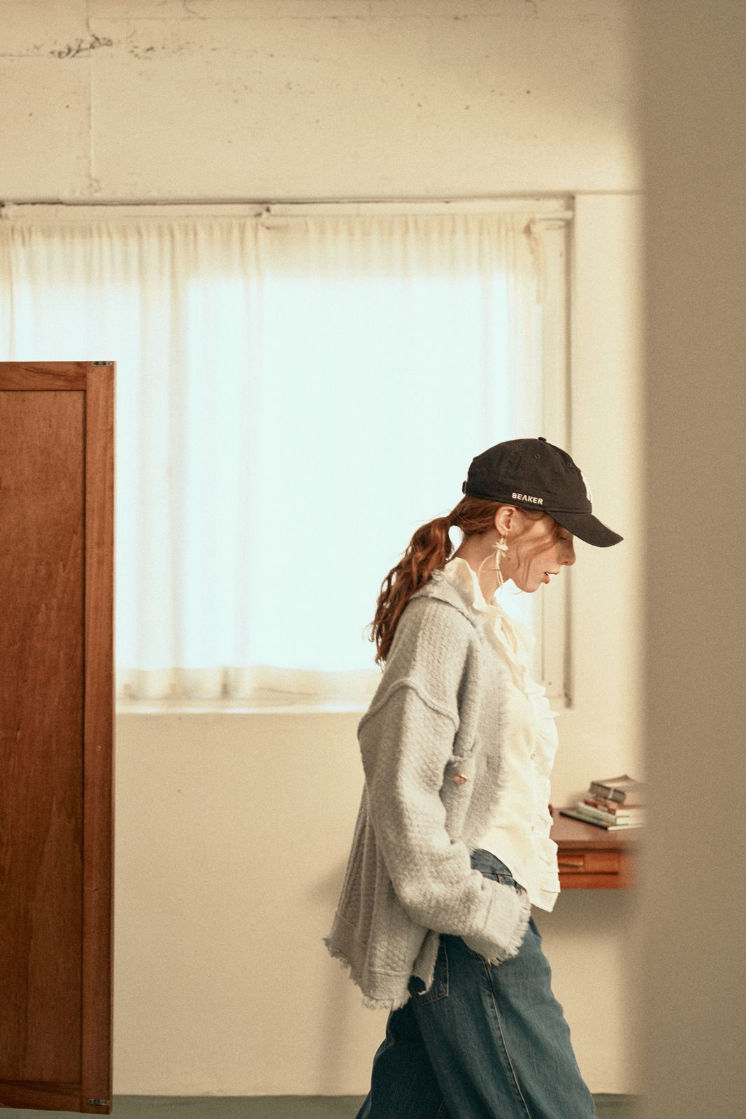 Opendoor · 패션 — 원목 문 옆을 지나는 옆모습 워킹 컷, 블랙 캡과 그레이 가디건, 화이트 러플 블라우스, 데님 팬츠 착용
