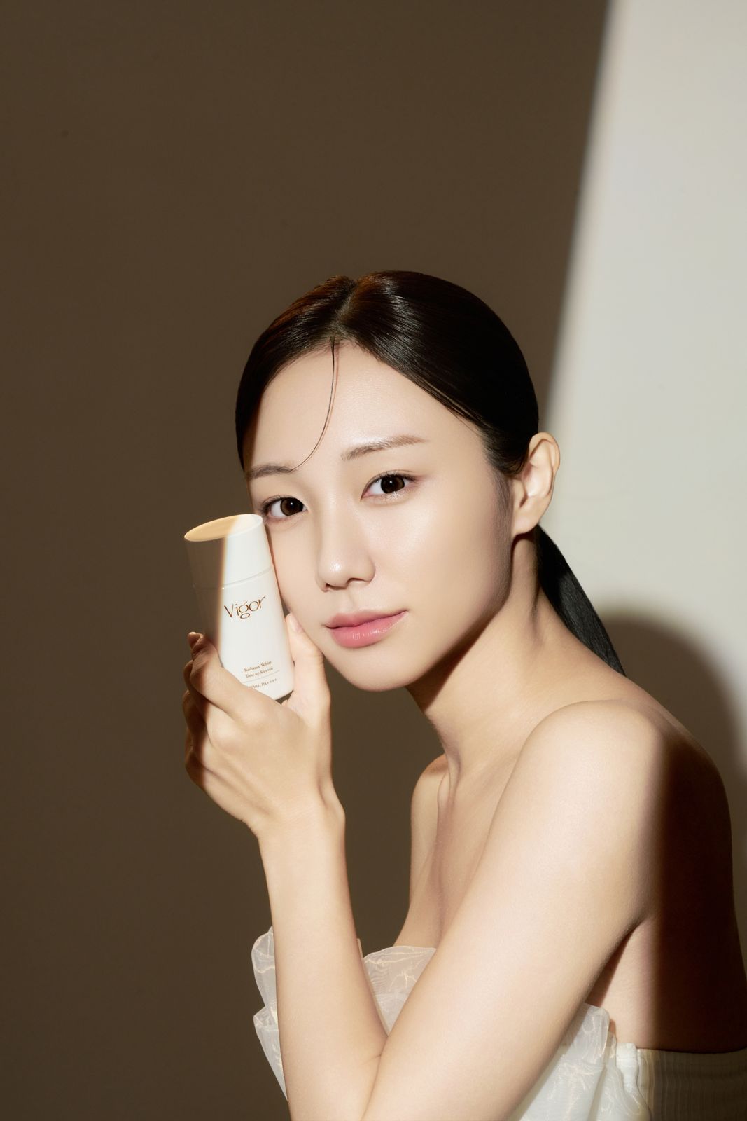 VIGOR · 선크림 (모델컷) — 대각선 강한 그림자와 웜톤 배경 속 선크림 튜브를 뺨에 댄 무드 모델컷